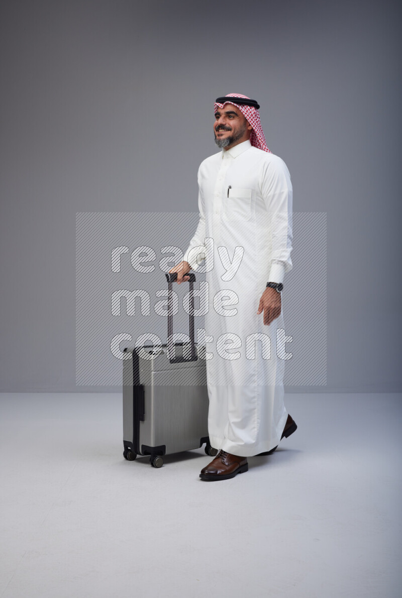Saudi man wearing Thob and red Shomag standing holding Travel bag on Gray background
