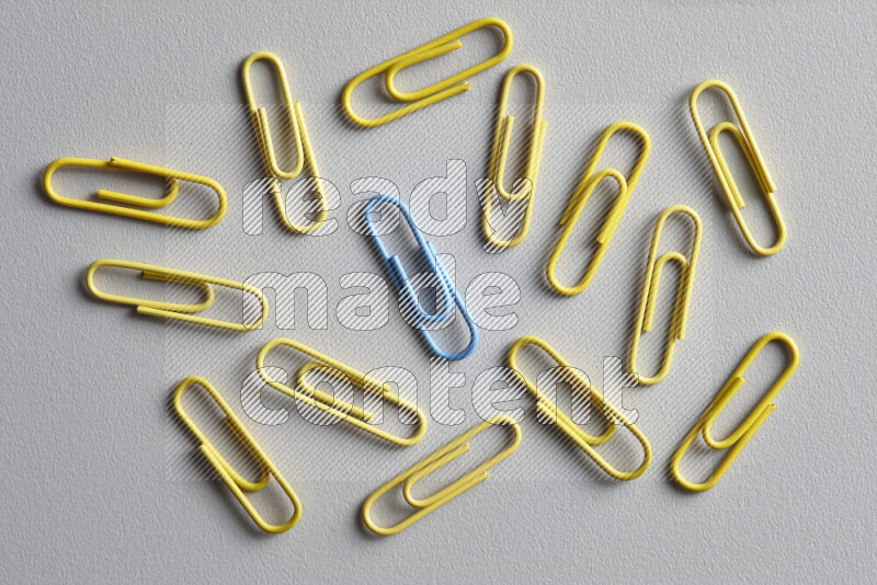 A blue paperclip surrounded by bunch of yellow paperclips on grey background