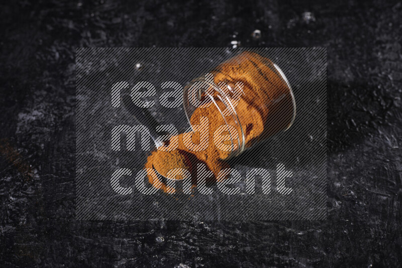 A glass jar full of ground paprika powder flipped with some spilling powder on black background