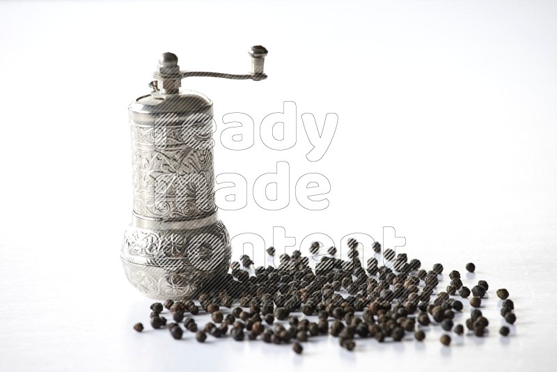 A turkish metal pepper grinder and spreaded black pepper beads on a white flooring