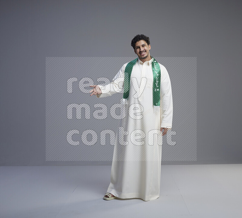 A Saudi man standing wearing thob and Saudi flag scarf on gray background