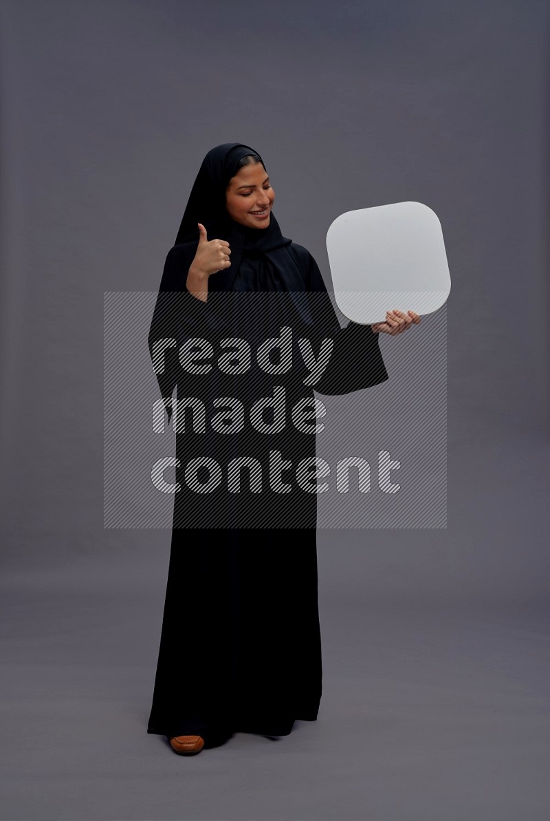 Saudi woman wearing Abaya standing holding social media sign on gray background
