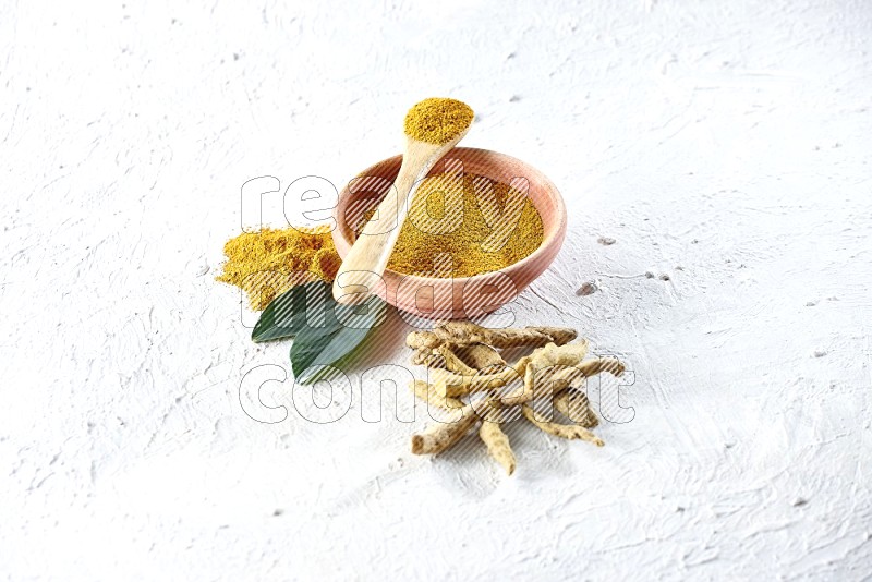 A wooden bowl and wooden spoon full of turmeric powder with dried turmeric fingers beside it on textured white flooring