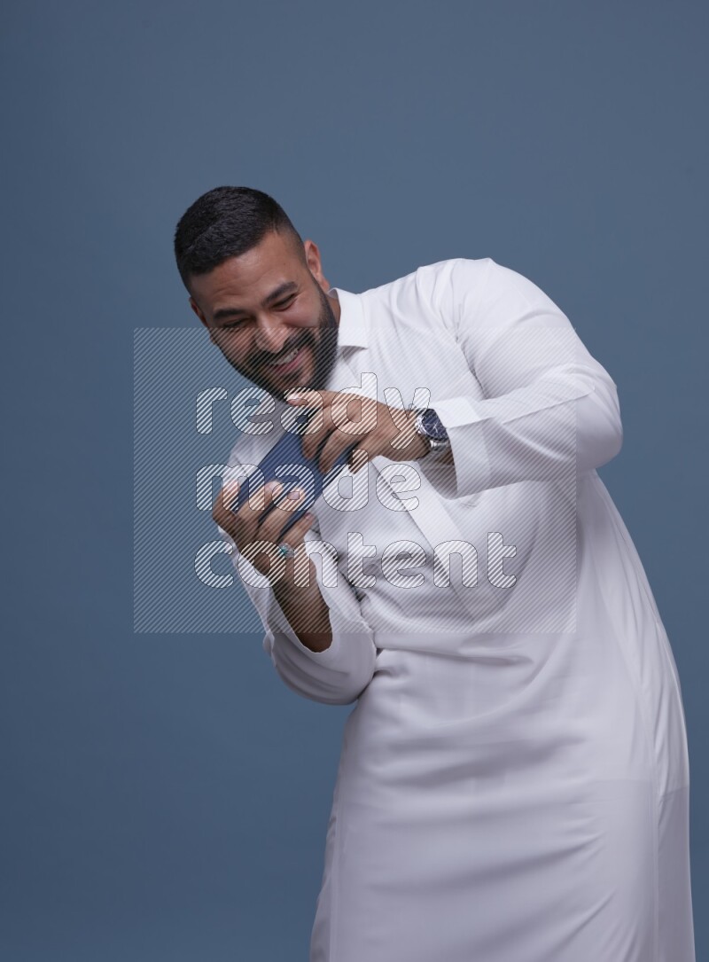 A man Playing a Game on his Smartphone  on Blue Background wearing Saudi Thob
