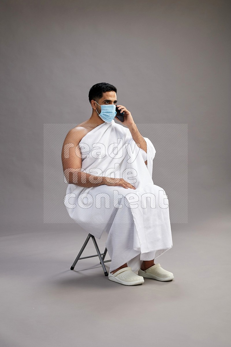 A man wearing Ehram with face mask sitting on chair talking on phone on gray background