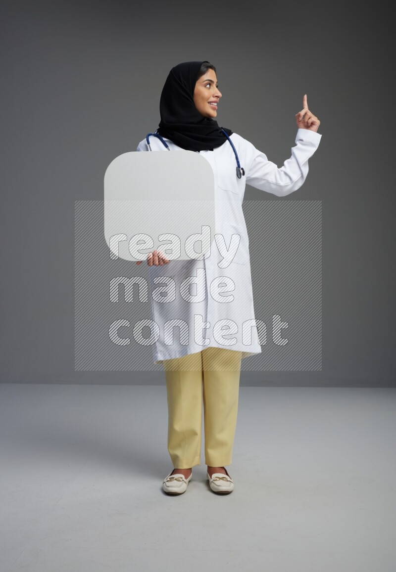 Saudi woman wearing lab coat with stethoscope standing holding social media sign on Gray background