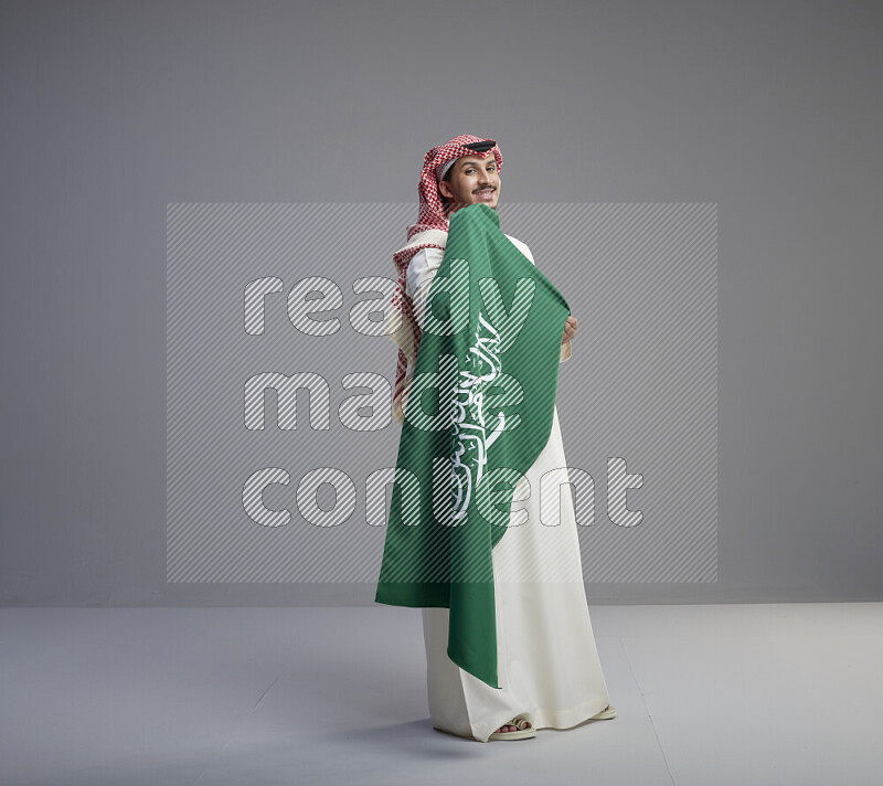 A saudi man standing wearing thob and red shomag holding big saudi flag on gray background