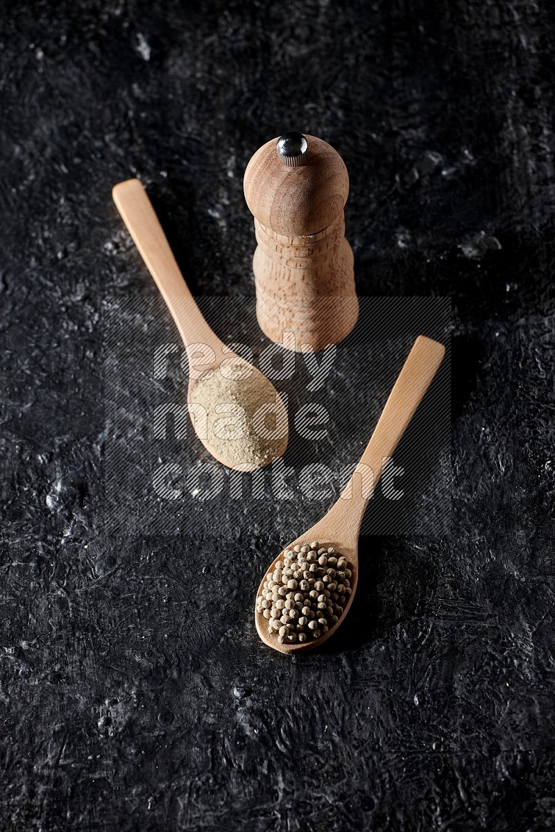 2 wooden spoons one full of white pepper powder and the other with pepper beads and a wooden pepper mill on textured black flooring