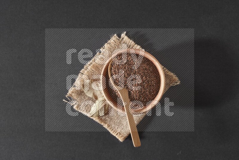 A wooden bowl full of flaxseeds with wooden spoon full of the seeds on it on burlap fabric on a black flooring