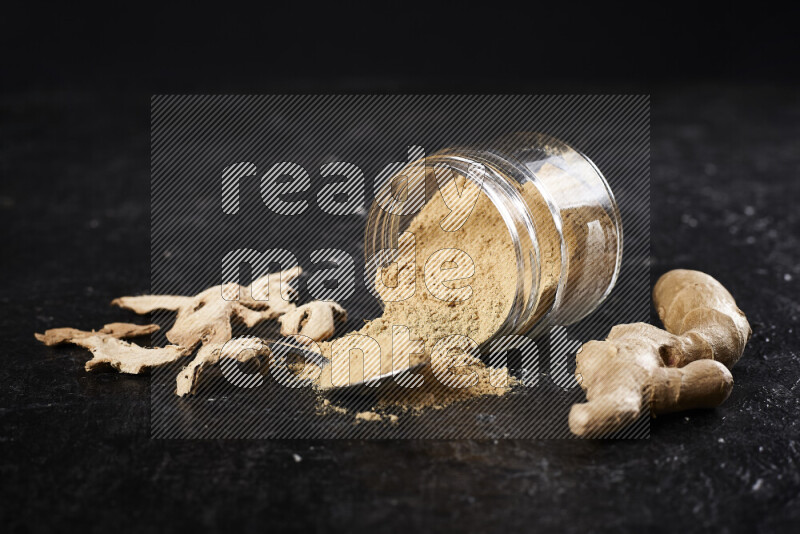 A glass jar full of ground ginger powder flipped with some spilling powder on black background