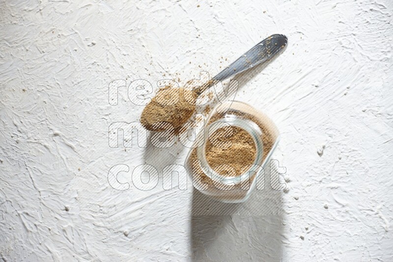 A glass spice jar and metal spoon full of cumin powder on textured white flooring