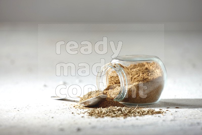 A glass spice jar and metal spoon full of cumin powder and the jar flipped and powder spilled out with cumin seeds on textured white flooring