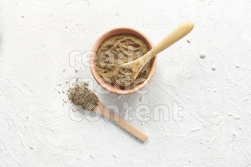 A wooden bowl and 2 wooden spoons full of cumin powder and cumin seeds on textured white flooring