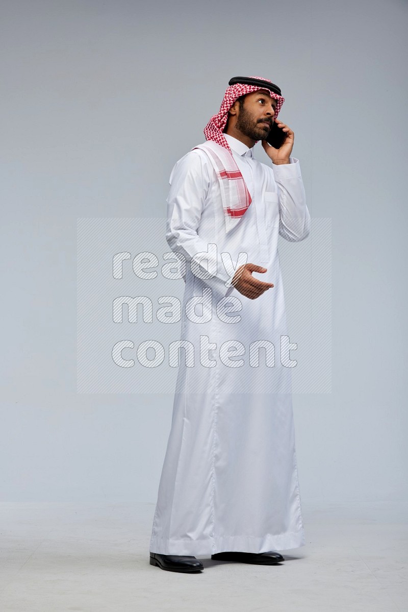 Saudi man Wearing Thob and shomag standing talking on phone on Gray background