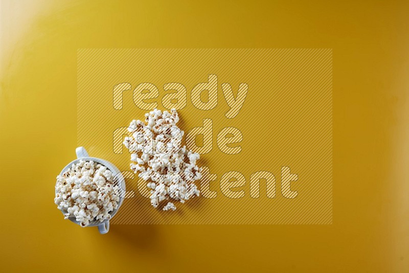 A white ceramic bowl full of popcorn with popcorn beside it on a yellow background in a top view shot