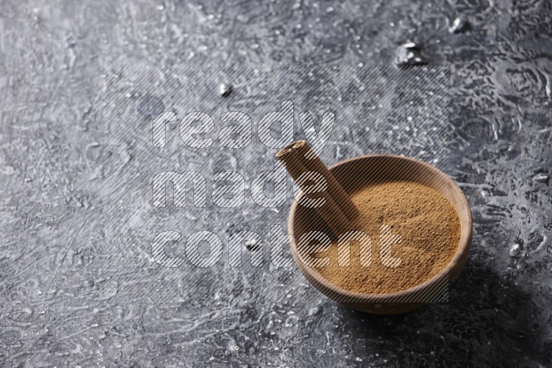 Wooden bowl full of cinnamon powder and a cinnamon stick on a textured black background