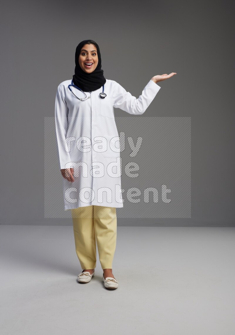 Saudi woman wearing lab coat with stethoscope standing interacting with the camera on Gray background