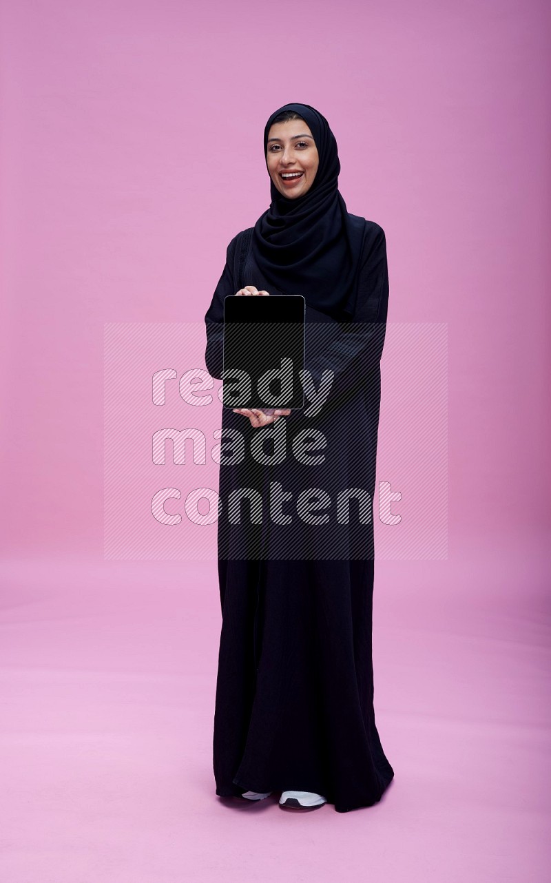 Saudi woman wearing Abaya standing showing tablet to camera on pink background