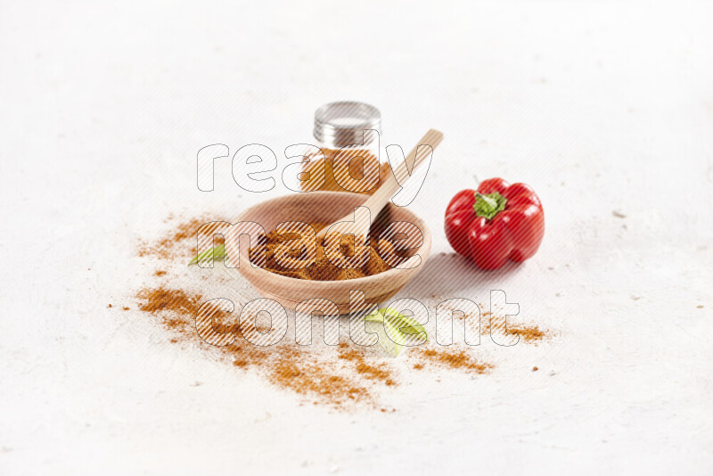 A wooden bowl full of ground paprika powder with a glass jar beside it and red bell pepper on white background