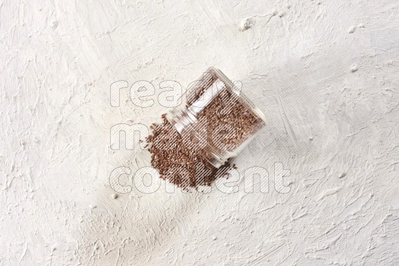 A glass jar full of flax seeds flipped and seeds spread out on a textured white flooring