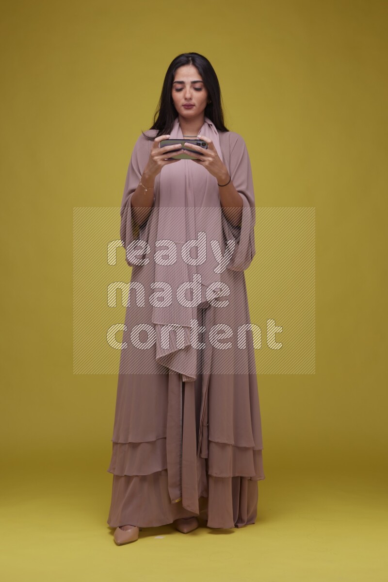 A woman Playing a Game on her smartphone on a Yellow Background wearing Brown Abaya