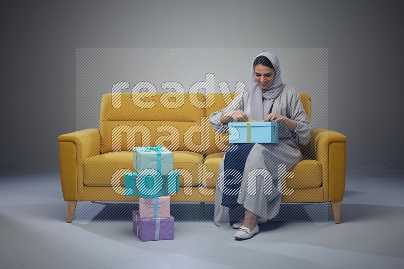 A Saudi woman wearing a light gray Abaya and head scarf sitting on a yellow sofa holding a gift box beside multi color gift boxes eye level on a grey background