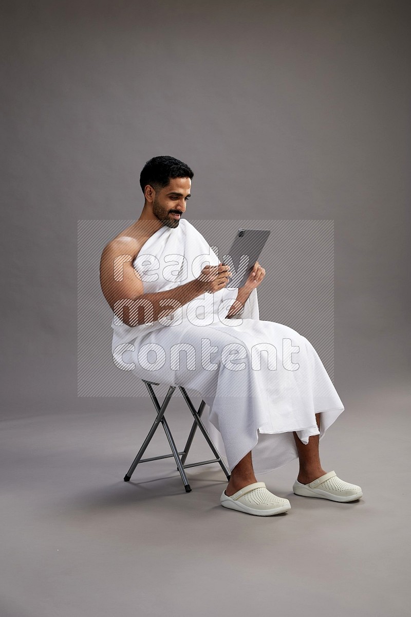 A man wearing Ehram sitting on chair holding a tablet on gray background