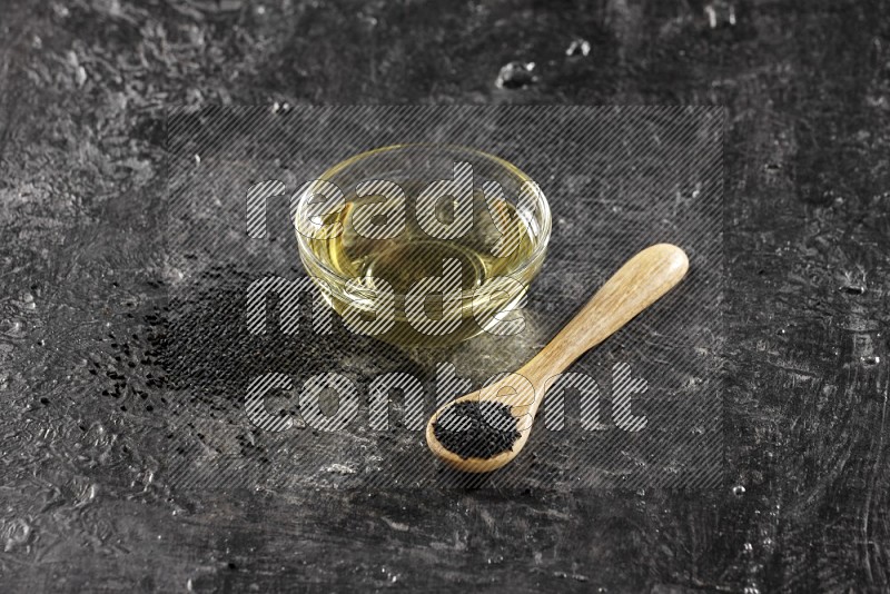 وعاء زجاجي ممتلئ بزيت حبة البركة وملعقة خشبية ممتلئة بالبذور مع بعض البذور متناثرة على أرضية سوداء