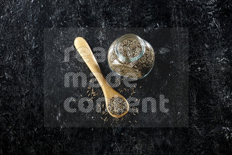 A glass spice jar and a wooden spoon full of cumin seeds on a textured black flooring