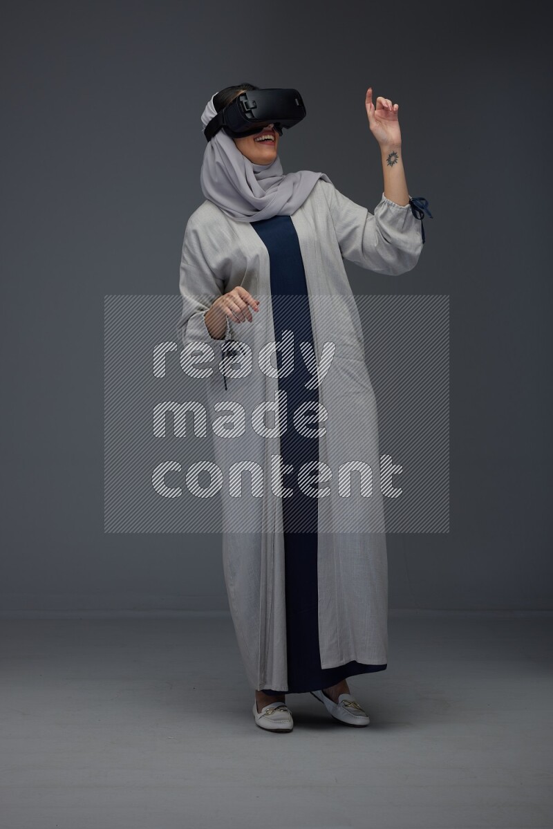 A Saudi woman wearing a light gray Abaya and head scarf standing and wearing VR eye level on a grey background