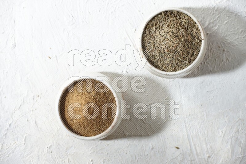 2 beige bowls full of cumin seeds and powder on a textured white flooring