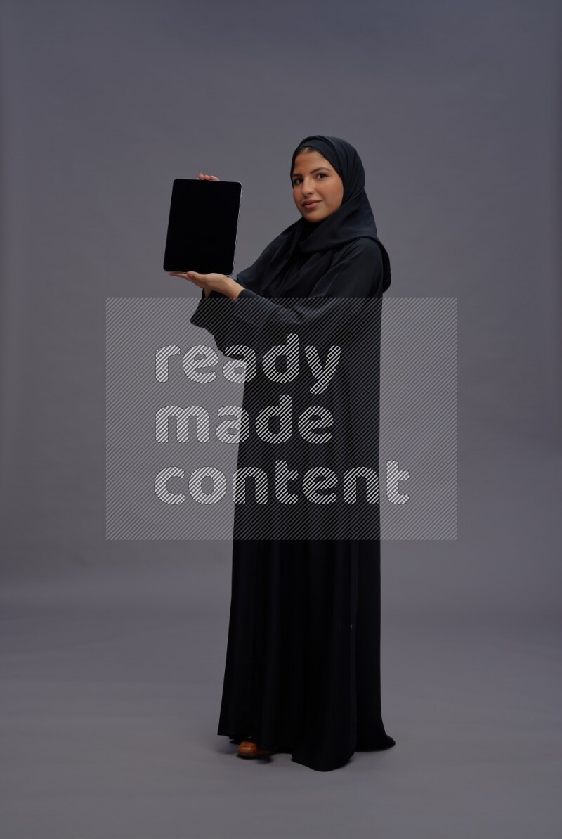 Saudi woman wearing Abaya standing showing tablet to camera on gray background