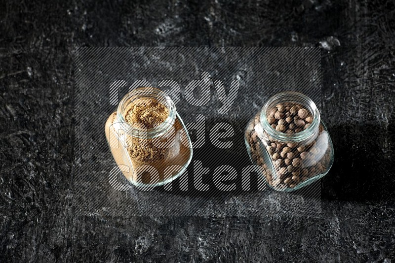 2 glass spice jars full of allspice powder and whole balls on a textured black flooring