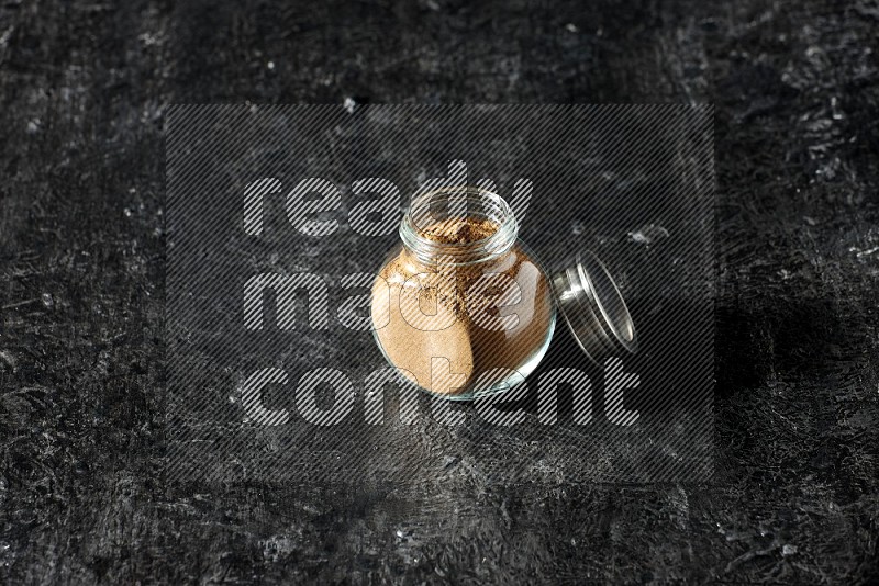 A glass spice jar full of allspice powder on a textured black flooring