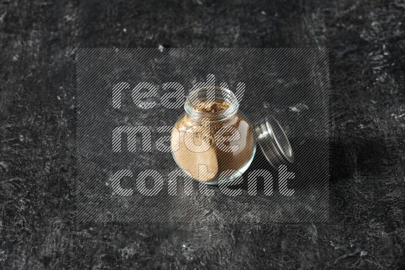 A glass spice jar full of allspice powder on a textured black flooring