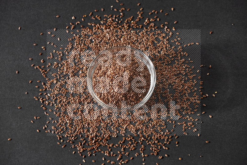 A glass bowl full of flaxseeds surrounded by the seeds on a black flooring
