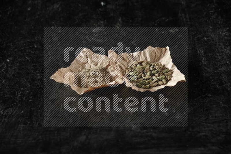 Cardamom seeds and cardamom powder in 2 crumpled pieces of paper on textured black flooring