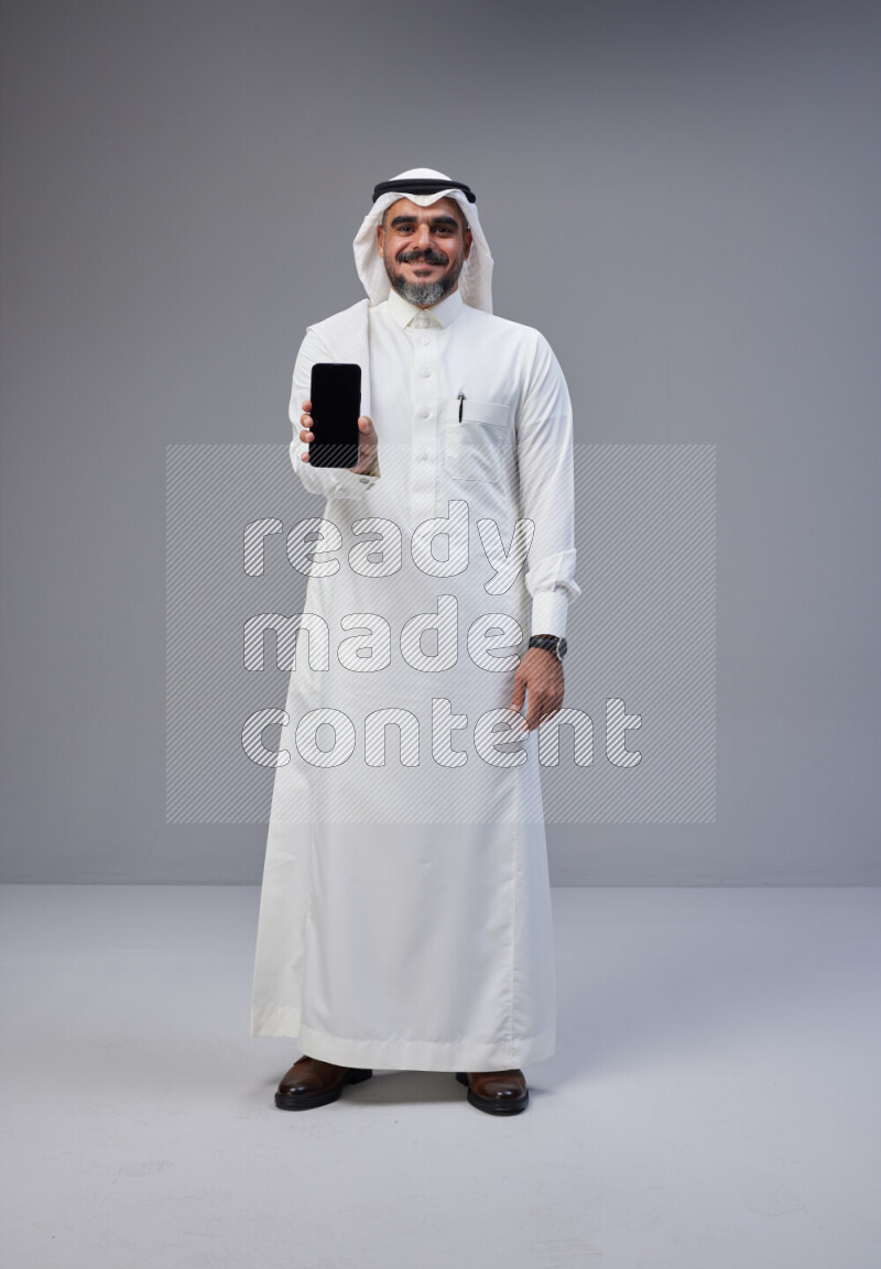 Saudi man Wearing Thob and white Shomag standing showing phone to camera on Gray background