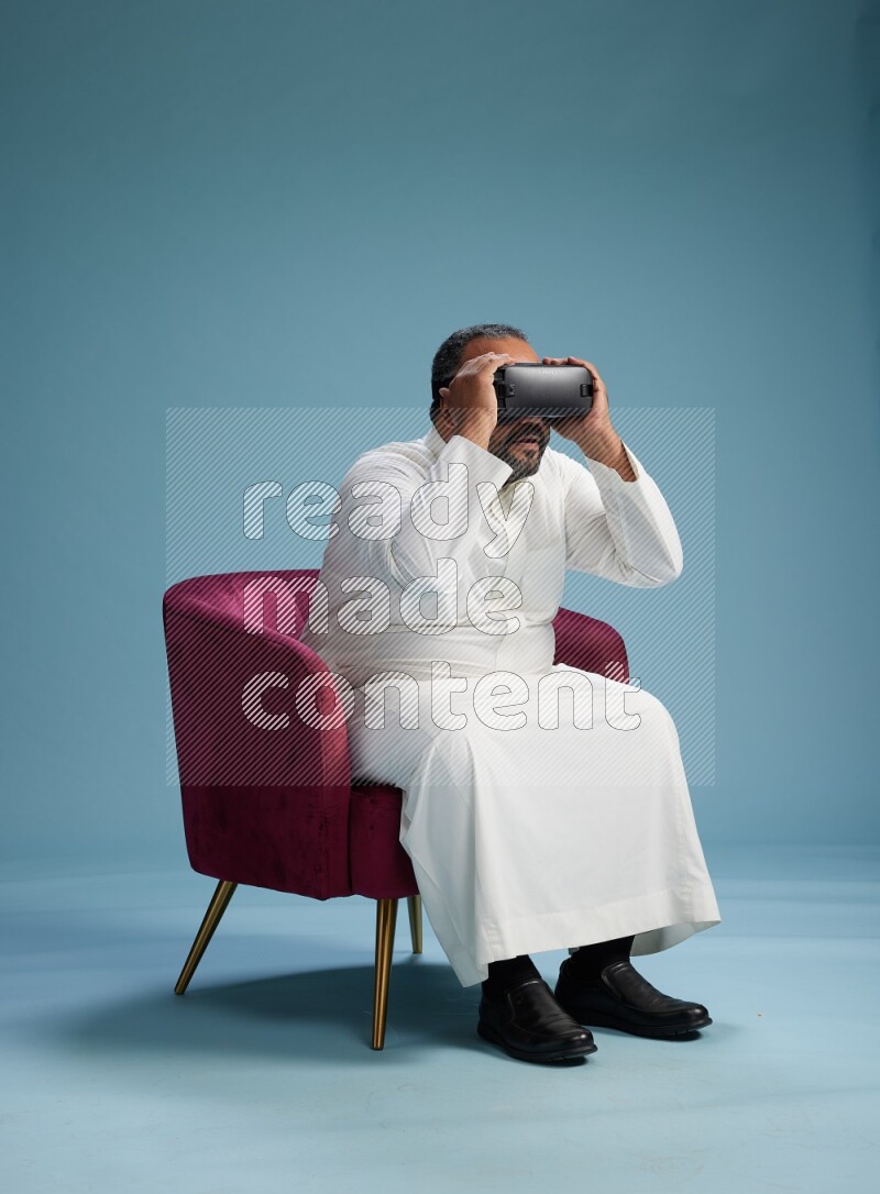 Saudi Man without shimag sitting on chair  gaming with VR on blue background