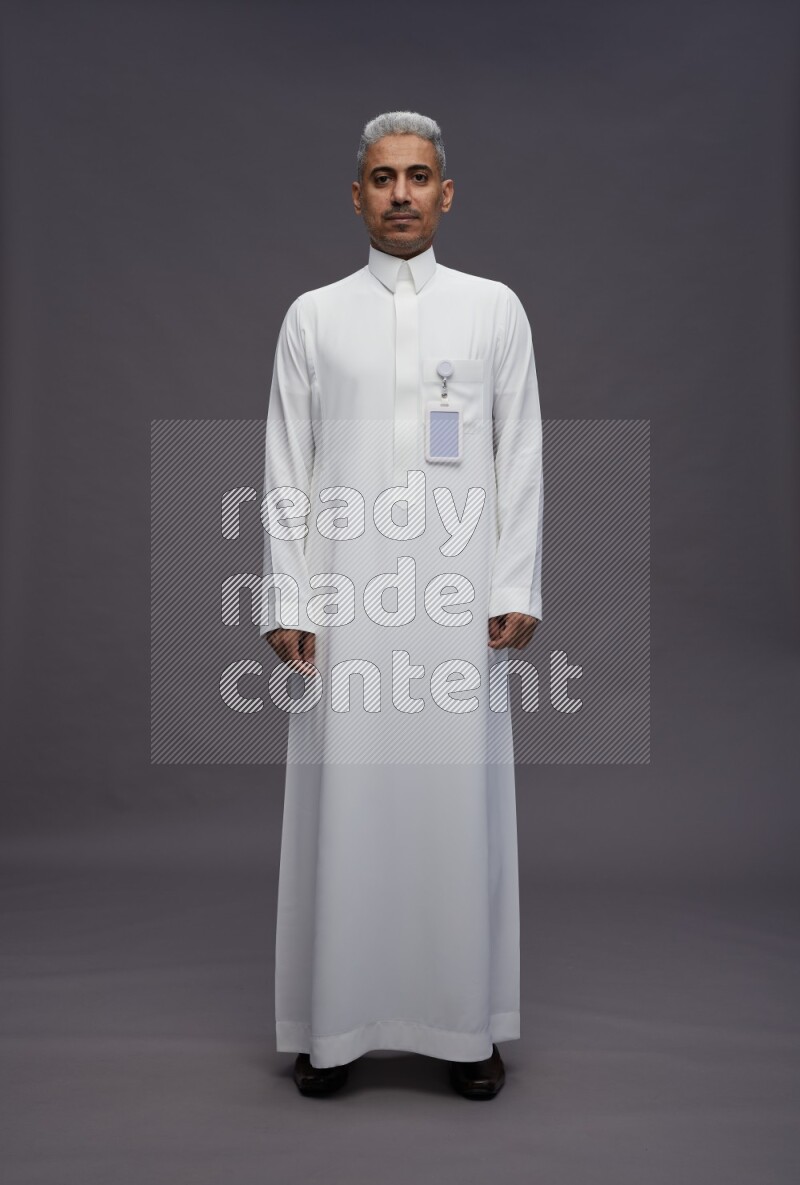Saudi man wearing thob with pocket employee badge standing interacting with the camera on gray background