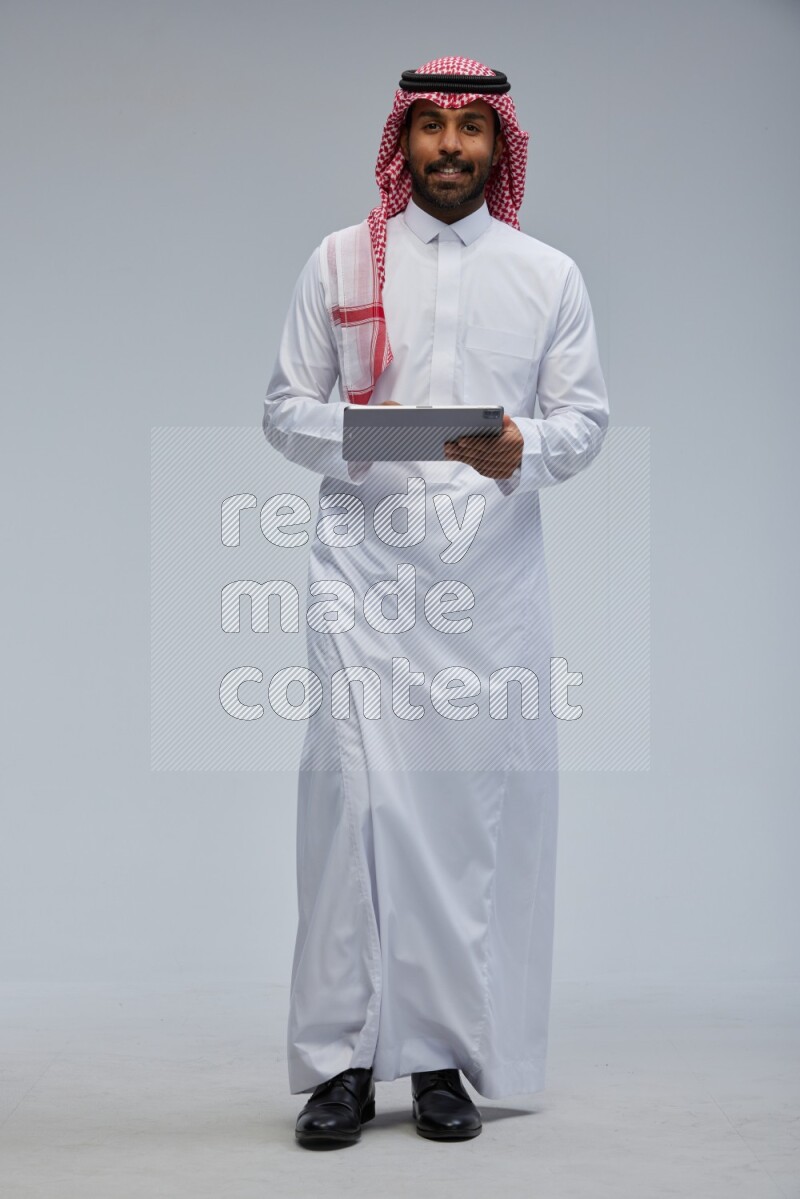 Saudi man Wearing Thob and shomag standing working on tablet on Gray background
