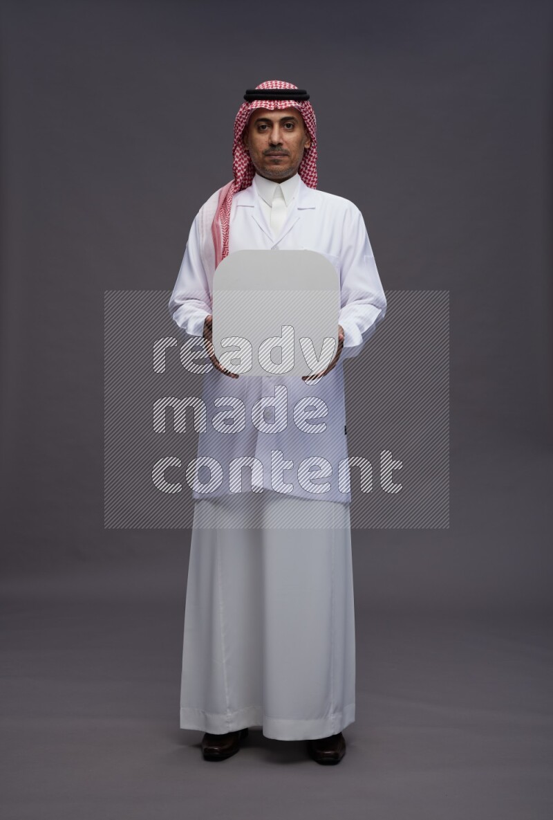 Saudi man wearing thob with lab coat and shomag with pocket employee badge standing holding social media sign on gray background