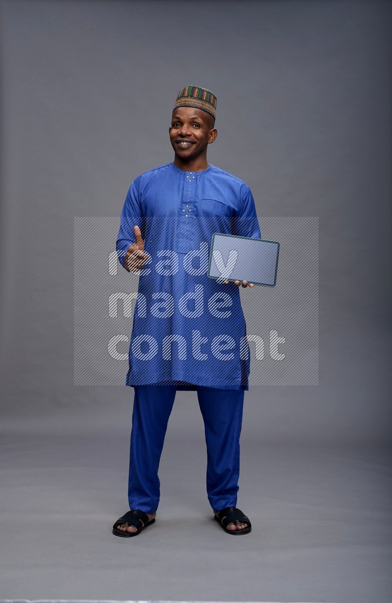 Man wearing Nigerian outfit standing showing tablet to camera on gray background