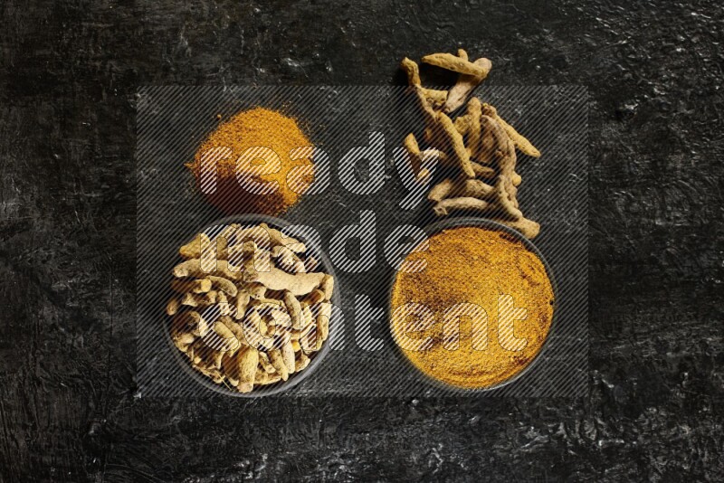 2 pottery black bowls, one full of turmeric powder and the other full of dried turmeric whole fingers on a textured black flooring