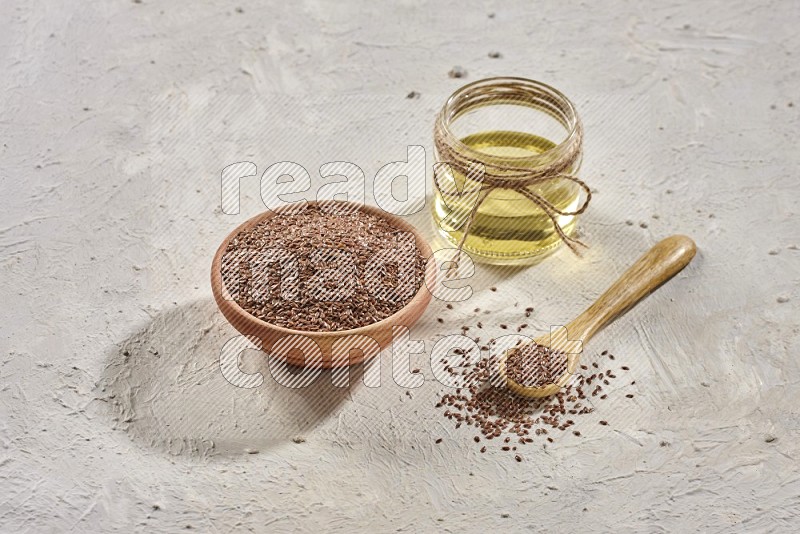 A wooden bowl and spoon full of flax and a glass jar of flax oil on a textured white flooring in different angles