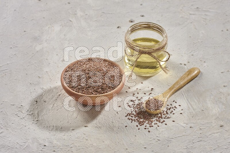 A wooden bowl and spoon full of flax seeds and a glass jar of flaxseeds oil on a textured white flooring