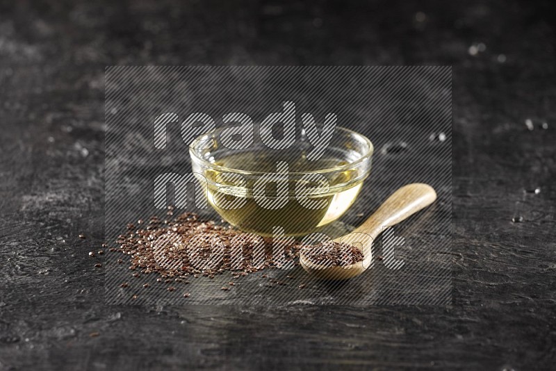 A glass bowl full of flax oil and wooden spoon full of flax with seeds spreaded on a textured black flooring in different angles