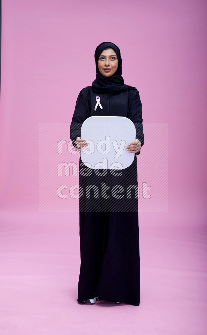 Saudi woman wearing pink ribbon on Abaya standing holding social media on pink background