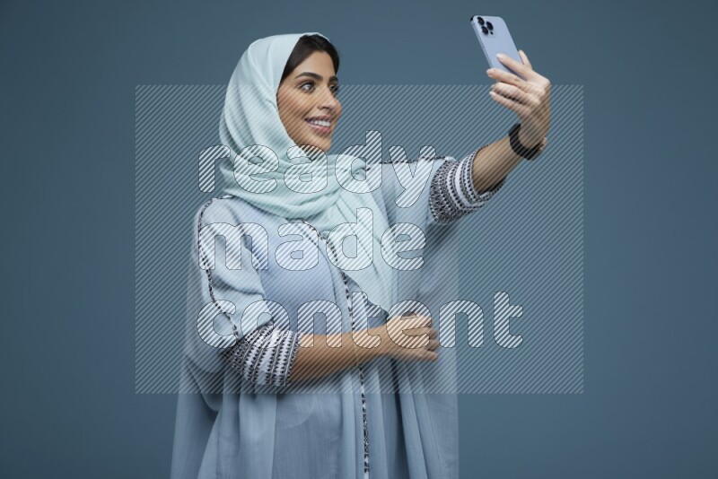A Saudi woman Taking a Selfie on a blue background wearing a blue Abaya with hijab