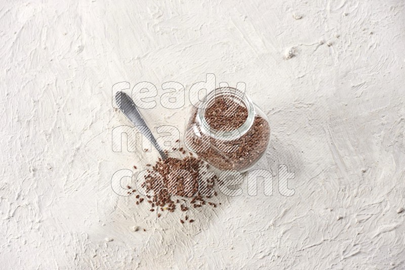 A glass spice jar full of flax seeds and a metal spoon full of the seeds on a textured white flooring
